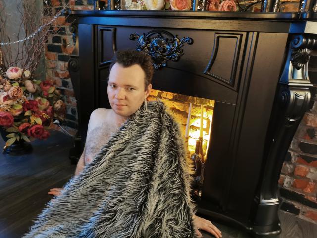 GenrikhBecca, shirtless with a flaccid chest, sits in front of a black fireplace, covered in a furry grey blanket, looking at the camera. Fireplace is lit, roses in background.