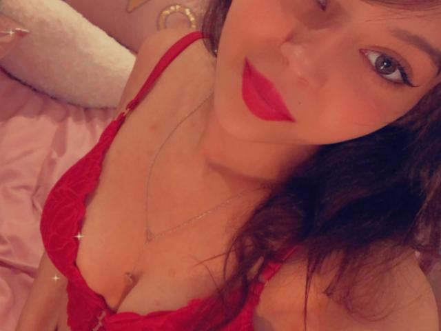 Bellefrancaise69 con un sujetador rojo de encaje, mostrando sus pechos, con lápiz labial rojo y ojos azules, acostada en una cama rosa, llevando un collar, con cabello castaño largo..