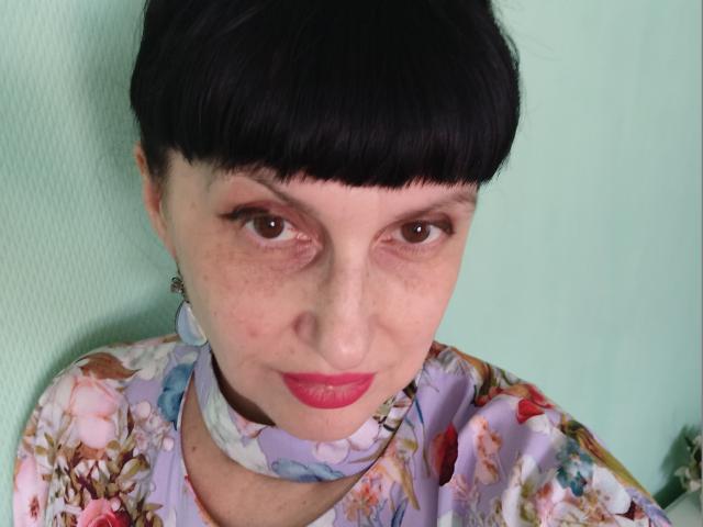 MelanyBanks has short black hair, freckled skin, and red lipstick. She's wearing a floral dress with a matching scarf, and has a choker necklace. Her eyes are brown.