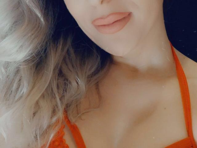 SophieDeee's face with slightly parted pink lips, light skin, and wavy blonde hair. She's wearing an orange lace bra, showing her cleavage. Soft light highlights her smooth skin.