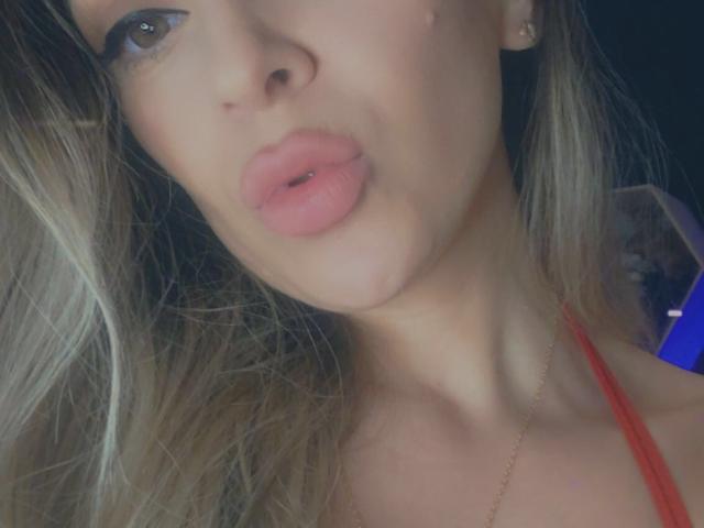 SophieDeee's face with pouty pink lips, slightly parted, showing her upper chest and blonde hair. She's wearing a red top with a gold necklace. Soft lighting.