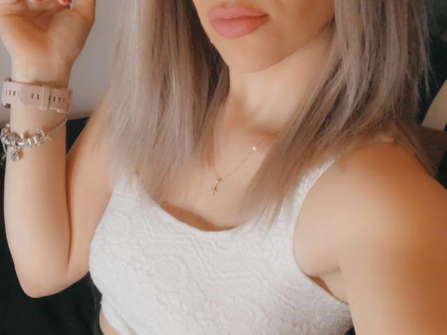 SophieDeee has blonde hair, pink lips, and wears a white lace tank top, a pink smartwatch, and a silver bracelet with a cross necklace. Boobs visible.