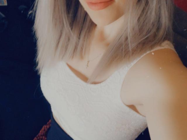 SophieDeee lleva un lápiz labial rosa claro y tiene la piel pálida. Viste una blusa blanca, sin mangas y con un estampado de encaje, que deja ver sus pequeños senos. También lleva un delicado collar de cruz de oro..