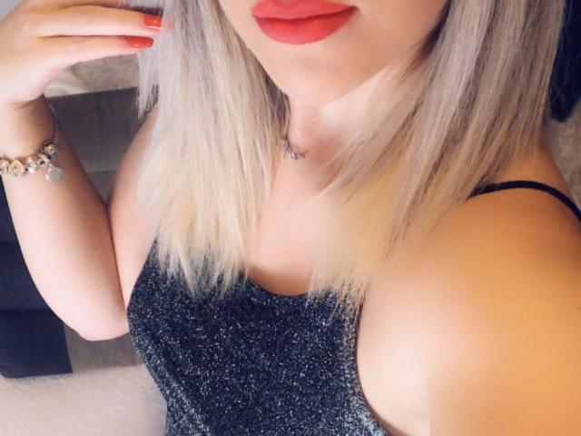 SophieDeee with blonde hair, red lipstick, sparkling black dress, exposed shoulder, red nail polish, and bracelet, showing off her tits and ass.