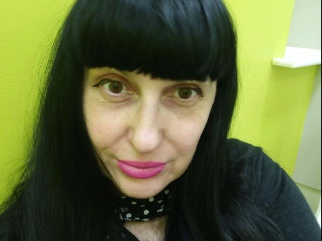 MelanyBanks has black hair with bangs, pink lipstick, and brown eyes, wearing a black polka dot scarf, smiling slightly, with a yellow-green background.