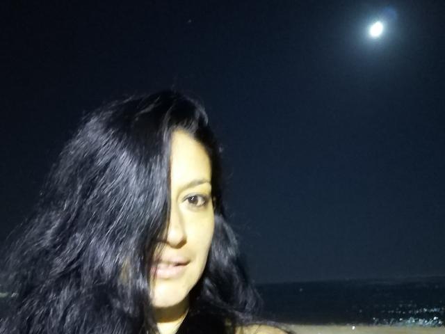 WetSweetKim mit dunklem, gewelltem Haar, das ein Auge bedeckt, blickt unter einem hellen Mond in einem Nachthimmel in die Kamera. Ihr Gesicht ist leicht beleuchtet..