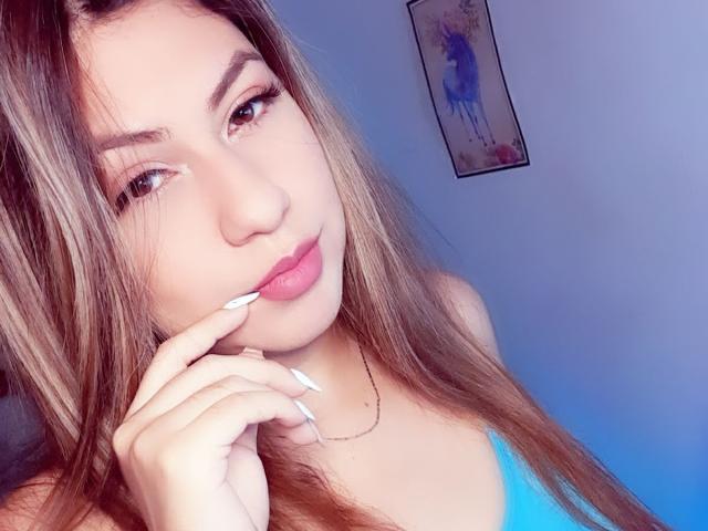 AnastaciaMonaco with long brown hair, pink lips, and blue top, touching her chin with white-manicured fingers, wearing a gold necklace, looking at the camera.