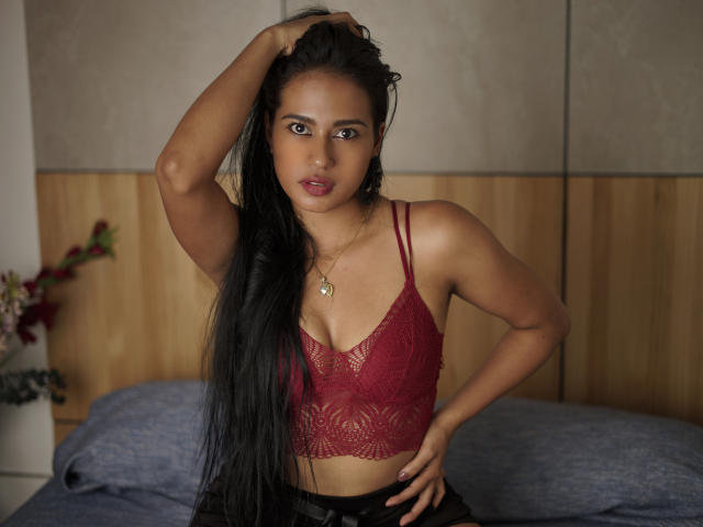 Thalia Cohen, com cabelos pretos longos, sutiã de renda vermelho, seios pequenos, pele bronzeada, mão no cabelo, sentada na cama, olhos escuros, batom vermelho, colar de ouro, olhando para a câmera..