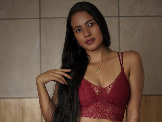 Thalia Cohen mit langem, schwarzem Haar, rotem Spitzen-BH, mittlerer Brustgröße, gebräunter Haut, einem leichten Lächeln, einer Hand im Haar, einer goldenen Halskette, braunen Augen, weicher Beleuchtung, dezentem Make-up und einer entspannten Pose..