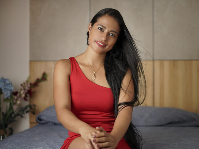 Thalia Cohen, con cabello negro largo, vestido rojo y collar, sentada en una cama, sonriendo, con las manos en el regazo, almohadas azules, cabecera de madera, arreglo floral en el fondo..