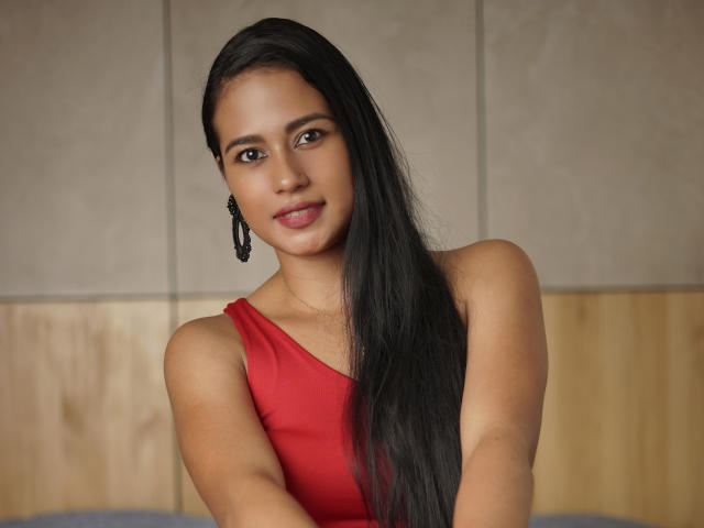 Thalia Cohen, con cabello negro largo, un vestido rojo, aretes negros y una ligera sonrisa, mostrando sus senos en una fotografía con iluminación suave..
