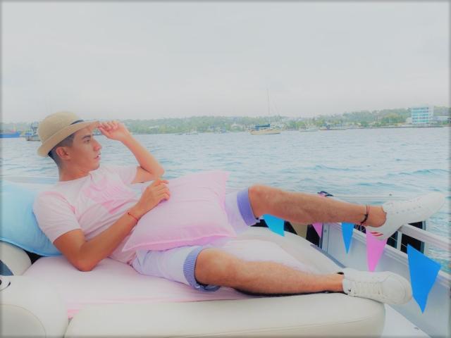 JordanSexyX, allongée sur un bateau, portant un chapeau beige, une chemise rose, un short blanc et des baskets blanches, avec un coussin rose, les jambes levées, regardant l'eau..