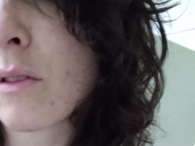 Il volto di LiveSex, con capelli scuri e disordinati che coprono parzialmente il naso e la bocca. Labbra rosa visibili e una piccola macchia scura sulla guancia sinistra. Pelle chiara..