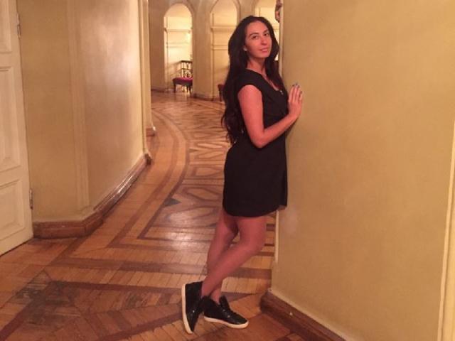 AsyaLu, con lunghi capelli scuri, un vestito nero e scarpe da ginnastica nere, in piedi contro un muro giallo, con le mani appoggiate al muro, che guarda indietro, in un corridoio con pavimento in legno..