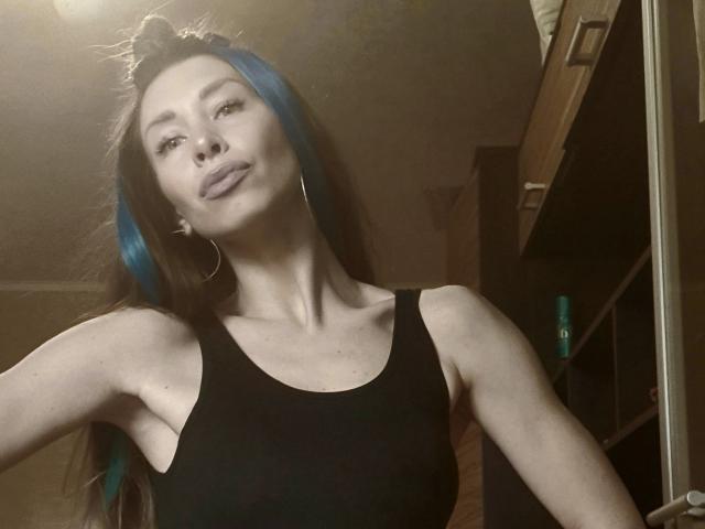 MisissFire with blue highlights in her hair, wearing a black tank top, pouting with her lips slightly parted, hoop earrings, and a top knot hairstyle, in a dimly lit room.