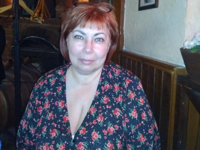 StellaDivafox mit roten Haaren, trägt ein schwarzes Kleid mit Blumenmuster, zeigt ihre Brüste in einem tief ausgeschnittenen Ausschnitt, lächelt und trägt einen Nasenring sowie dezentes Make-up..
