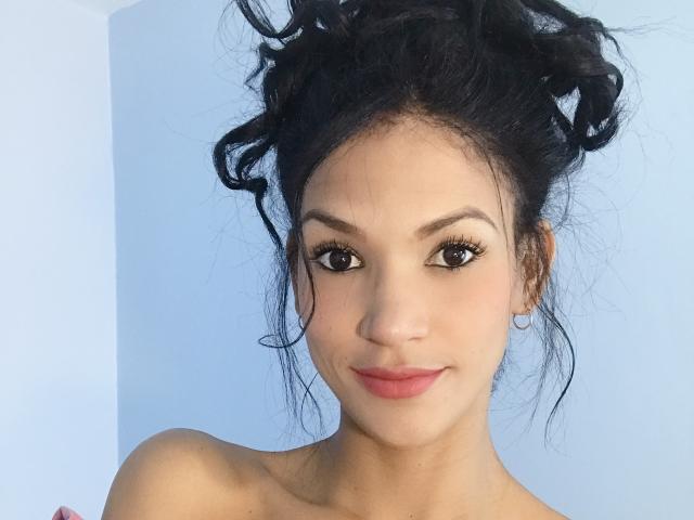 PaulinaNicolle with messy black hair in a bun, brown eyes, long lashes, pink lips, small hoop earrings, bare shoulders, light skin, subtle smile, looking at camera.