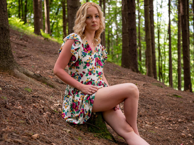 SoleildeNuit met blond haar, gekleed in een bloemenjurk, zittend in een bos, benen gekruist, waarbij haar dijen zichtbaar zijn, met een serieuze blik, handen op haar buik, dennennaalden op de grond..