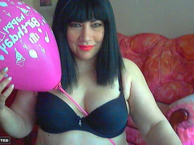 MelissaLight, con un sujetador negro, sosteniendo un globo rosa, tiene el cabello negro y liso, lápiz labial rojo y está sonriendo. Sus senos son de tamaño medio y está sentada en un sofá rosa..