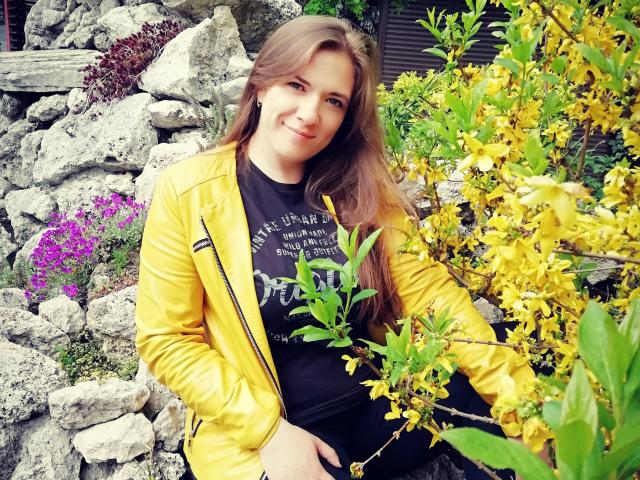 PatriciaSyde, com cabelos castanhos longos, jaqueta de couro amarela, camisa preta e um sorriso adorável, sentada ao lado de flores amarelas, mostrando uma leve barriga, com pedras ao fundo..