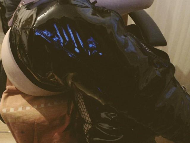 ChaudeOrgasme's shiny black latex pants are pulled down, exposing their round ass. They're wearing fishnet stockings and black boots, leaning forward. The image is a sexy, tight shot.