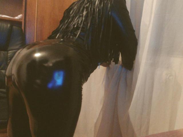 ChaudeOrgasme is bent over, wearing shiny black latex pants that highlight her ass. She has a black, fringed top and is looking through a white curtain.
