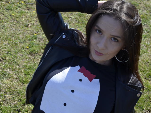AnaHathor with brown hair, black jacket, and white shirt with red bowtie, has small tits, hoop earrings, and is standing on grass, hand in hair, looking at camera.