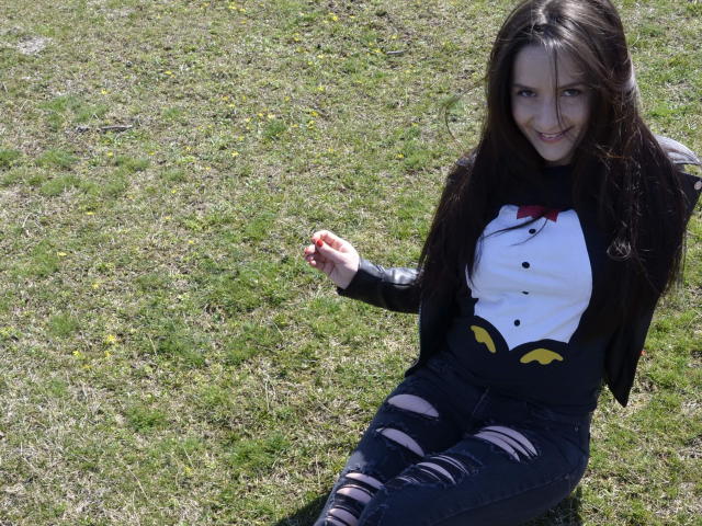 AnaHathor with long dark brown hair, smiling, wearing a black shirt with a white face print, black jacket, and ripped jeans, sitting on grass. Her tits are small.