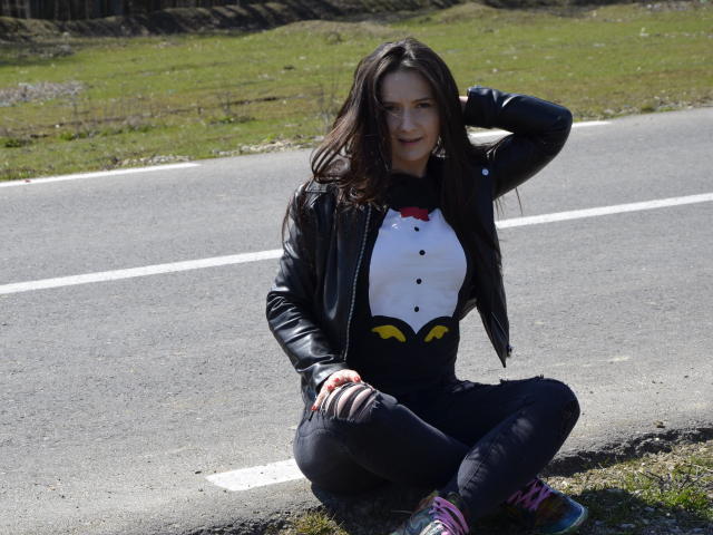 AnaHathor sits on a road, wearing a black leather jacket, white shirt with yellow and black patterns, black pants, and colorful sneakers. She has long brown hair and smiles.