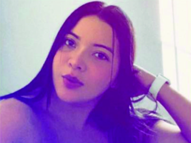 SamanthaWade, com cabelos longos e pretos, olhando para a câmera, com um sorriso discreto, usando uma pulseira branca, com iluminação suave em tons de roxo e azul..