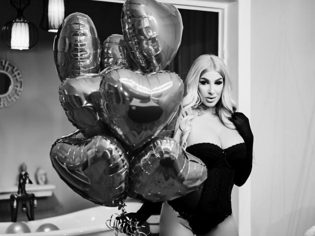 Black and white photo of TanyaMonroe with long blonde hair, large tits in a black lace corset, holding shiny silver heart balloons, standing in a bathroom.