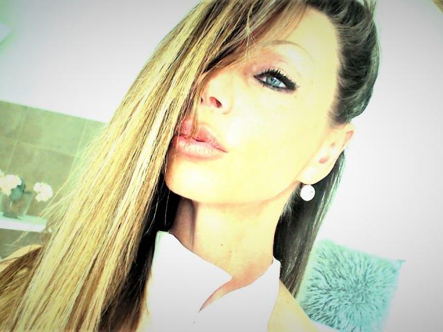 MonikaPrize has long, blonde hair covering one eye, blue eyeliner, and pink lips. She's wearing a white top and small, round earrings. Her face is slightly tilted.