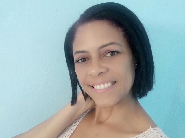 ErikaSpmt, com cabelo preto curto, sorrindo, segurando o pescoço, vestindo uma blusa branca de renda, mostrando seus seios pequenos e bonitos, e seus olhos castanhos pequenos e bonitos, em frente a uma parede azul..