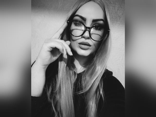 SerafimaArt with long blonde hair, thick eyebrows, round glasses, and full lips, touching her cheek with her finger, wearing a black top, in a black-and-white photo.