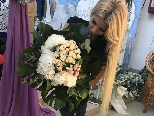 AlisaAleksandr, loira, com cabelos longos, segurando um buquê de rosas brancas e folhas verdes, sorrindo, vestindo uma blusa preta, em uma floricultura. Seios não visíveis..