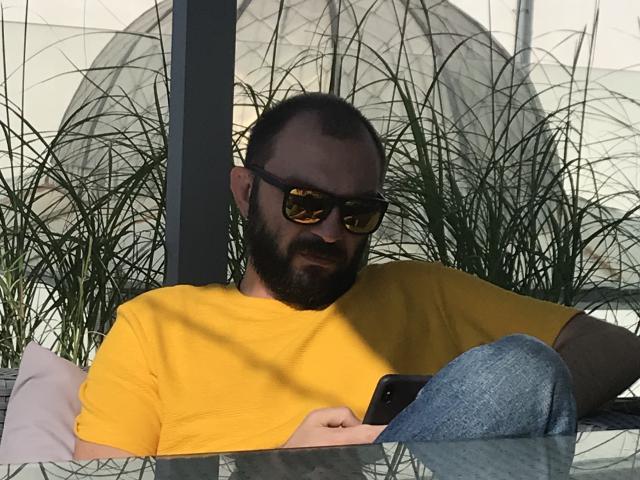 Ein Mann mit Bart, dunklen Sonnenbrillen, einem gelben Hemd und blauen Jeans, der ein Telefon hält und entspannt, leicht schmutzig aussehend, zurücklehnt, umgeben von grünen Pflanzenschatten..