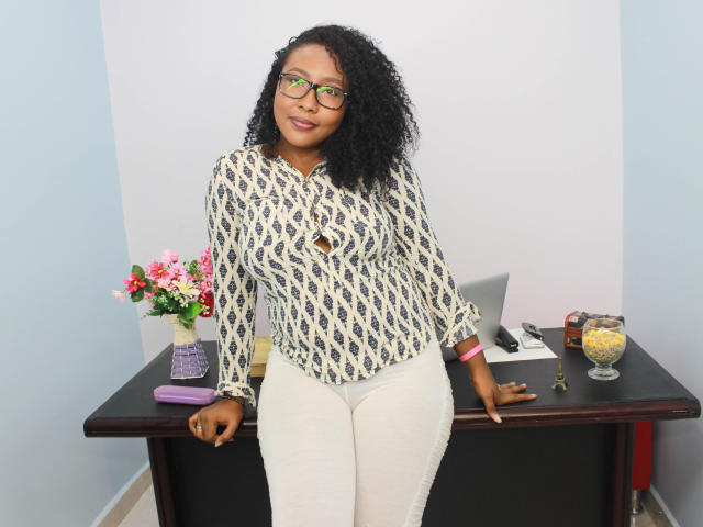 CaroldelaTorre with curly black hair, green glasses, patterned blouse, white pants, leaning on desk, flower vase, phone, glass with yellow drink, pink bracelet, smiling.