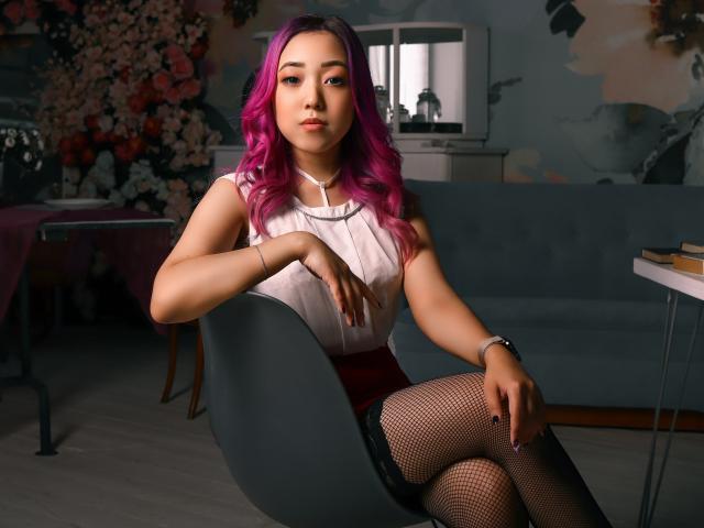 RenyLime con cabello morado, camiseta blanca sin mangas, falda roja, medias de rejilla y encaje negro, sentada en una silla gris, mirando a la cámara, con una mano en la rodilla, y un reloj en la muñeca derecha..