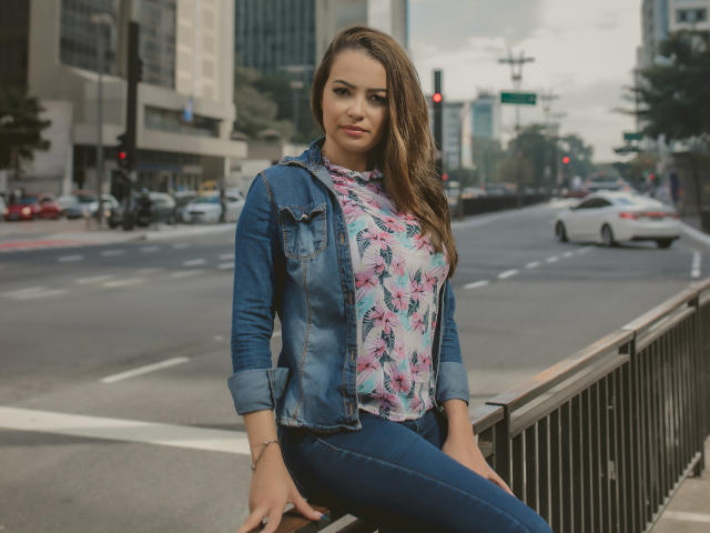 Amara, com longos cabelos castanhos, vestindo uma jaqueta jeans, uma blusa florida e calças jeans azuis, está em pé ao lado de um corrimão de metal em uma rua da cidade, com uma expressão confiante. Seus seios são visíveis..
