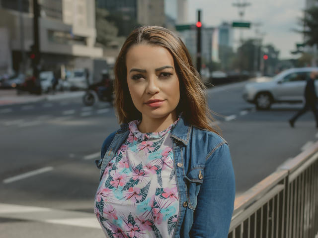 AmaraLove, avec une poitrine de taille moyenne, portant un haut fleuri et une veste en jean, debout dans une rue de la ville, cheveux bruns, maquillage discret, expression confiante, arrière-plan flou, environnement urbain..