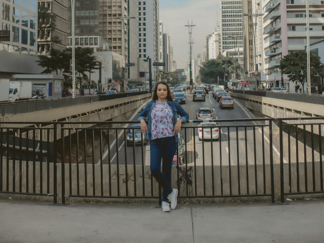 AmaraLove debout, les mains sur une clôture noire, portant une veste en jean, une chemise à fleurs, un jean bleu, des baskets blanches, tenant un vélo, rue urbaine avec des voitures en arrière-plan, lumière du jour, tenue décontractée..