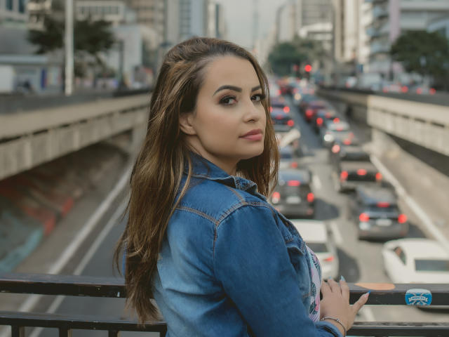 Amara, com longos cabelos castanhos, jaqueta jeans azul, olhando por cima do ombro, em pé em uma ponte, tráfego da cidade ao fundo, carros com luzes vermelhas e brancas, maquiagem discreta, expressão confiante..