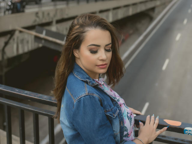 Amara, avec ses cheveux bruns, sa veste en jean bleu et son foulard floral rose, regardant vers le bas, tenant une rambarde, vue de profil, léger sourire, maquillage naturel, ongles bleus, journée nuageuse, arrière-plan urbain..