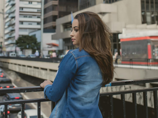 Amara, com cabelo castanho médio, vestindo uma jaqueta jeans azul, em pé perto de um corrimão preto, olhando para o lado, com um fundo de cidade com prédios e um ônibus vermelho..