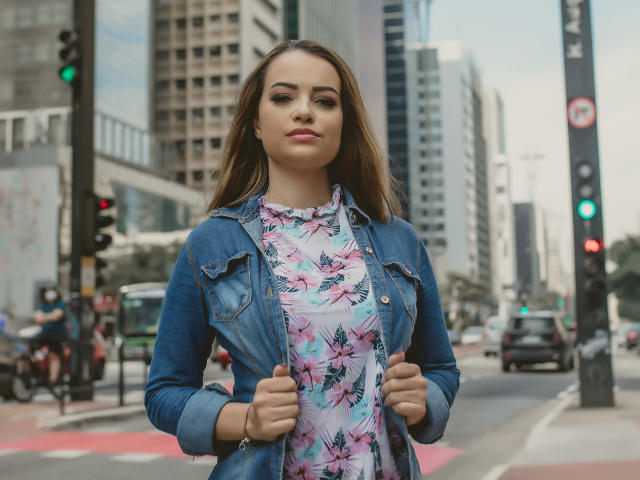 AmaraLove stands in a city street, wearing a denim jacket over a floral dress, hands gripping jacket edges, with a confident, sexy expression, brown hair, and subtle makeup.