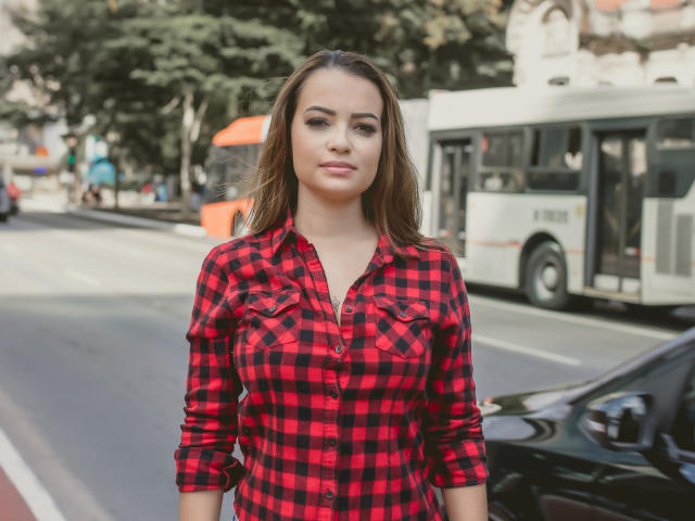 A AmaraLove tem cabelo castanho, veste uma camisa xadrez vermelha e preta, está em uma rua com um ônibus e um carro ao fundo, e parece confiante, com um leve sorriso..