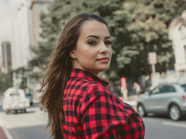 AmaraLove with long brown hair, red-black plaid shirt, looking over shoulder, soft smile, subtle makeup, city street background, blurred cars and buildings, sunny day, confident vibe.