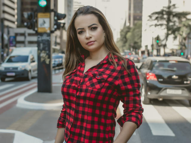 Amara, com seios médios, vestindo uma camisa xadrez vermelha e preta, está em uma rua da cidade com carros ao fundo, tem cabelo castanho e uma expressão confiante..