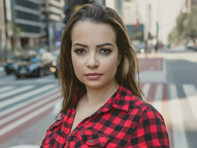 AmaraLove a les yeux marron, des lèvres pulpeuses et des cheveux bruns de longueur moyenne. Elle porte une chemise à carreaux rouge et noire, et se tient dans une rue de la ville, avec des voitures floues en arrière-plan..