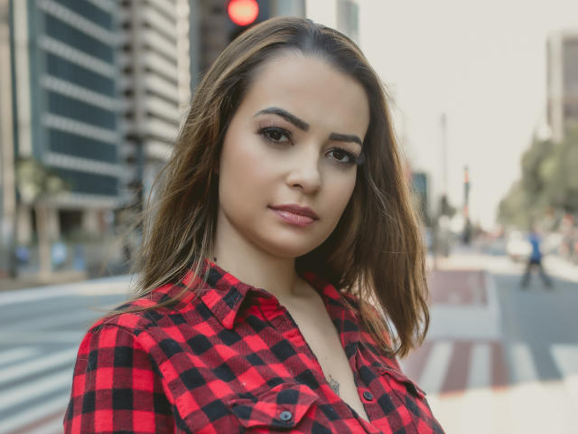 Amara, com cabelo castanho, camisa xadrez vermelha e preta, lábios cheios e maquiagem, em uma rua da cidade, mostrando levemente os seios, olhando para a câmera..